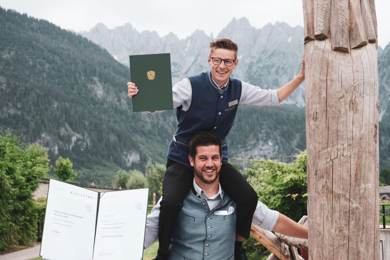 A man sits on the shoulders of another, both happily holding certificates towards the camera.