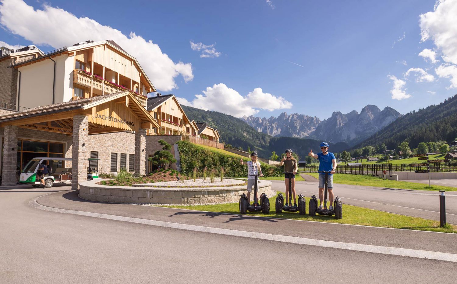 Drei Personen stehen mit Daumen hoch auf Segways, im Hintergrund das Dachsteinkönig Familux Resort.
