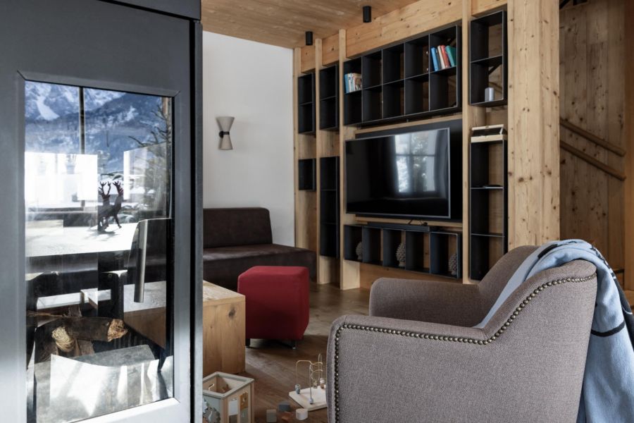 Gemütlicher Wohnraum mit Sessel vor einem Kamin, im Hintergrund eine Couch und ein TV an der Wand.