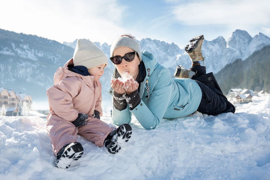 A man lying in the snow blowing snow into the camera, a small child sitting next to him.
